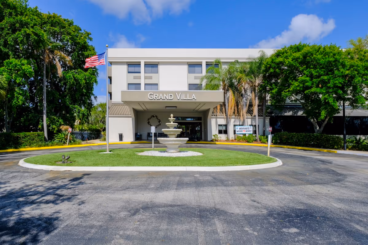 Front exterior view of the Grand Villa building with a circular driveway, a central fountain, an American flag on a flagpole, and surrounding trees under a blue sky with some clouds.