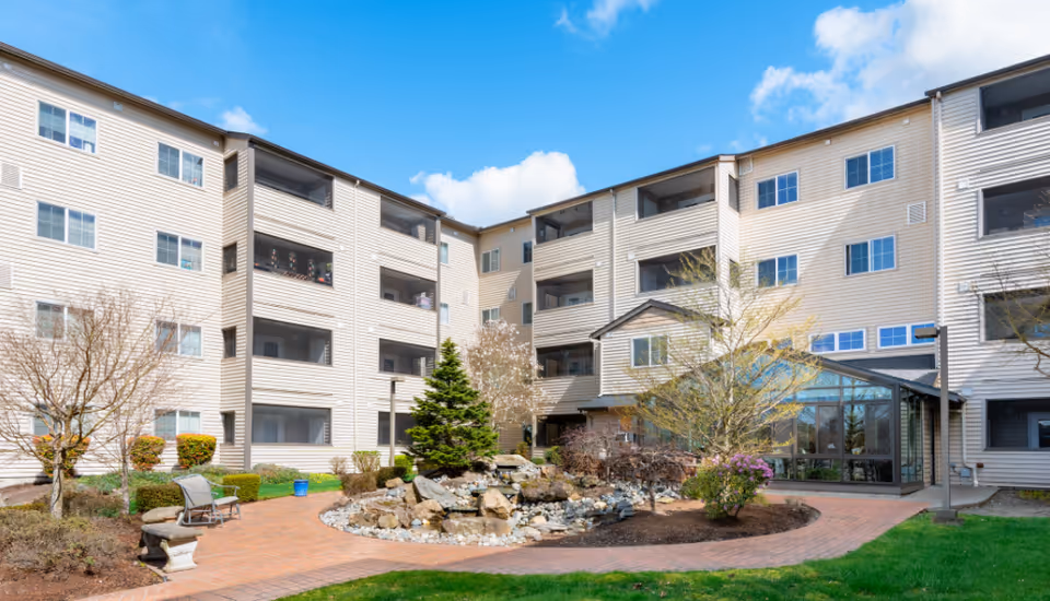 Exterior view of a multi-story senior living facility with beige siding, multiple windows, and screened balconies. In the foreground, there is a landscaped courtyard featuring a small rock water feature, trees, shrubs, benches, and a brick walkway under a blue sky with some clouds.