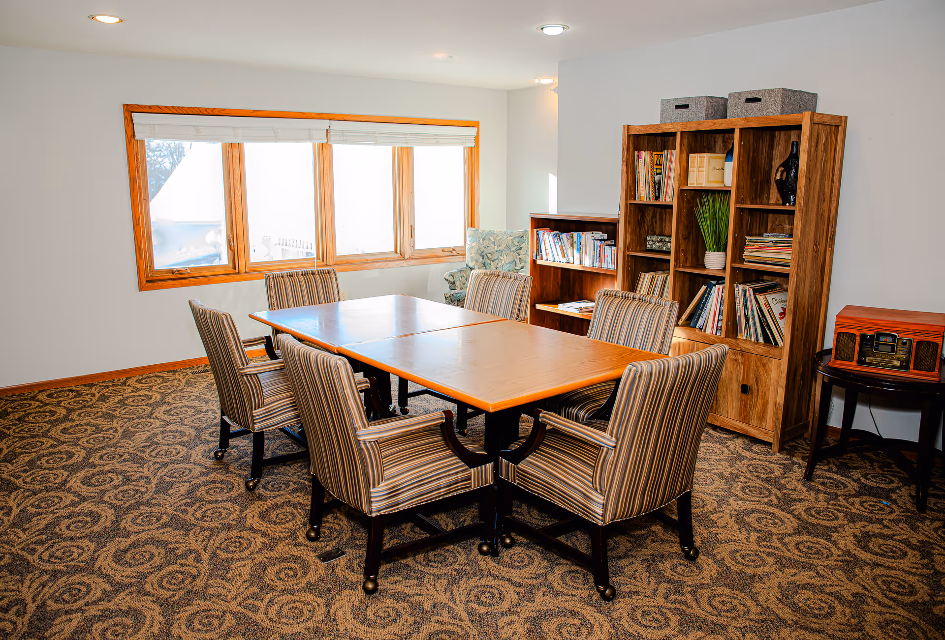 A cozy room with a wooden table surrounded by six striped upholstered chairs on a patterned carpet. There is a large window with white blinds letting in natural light. Against the wall, there are wooden bookshelves filled with books and decorative items, and a small table with a vintage-style radio on it.