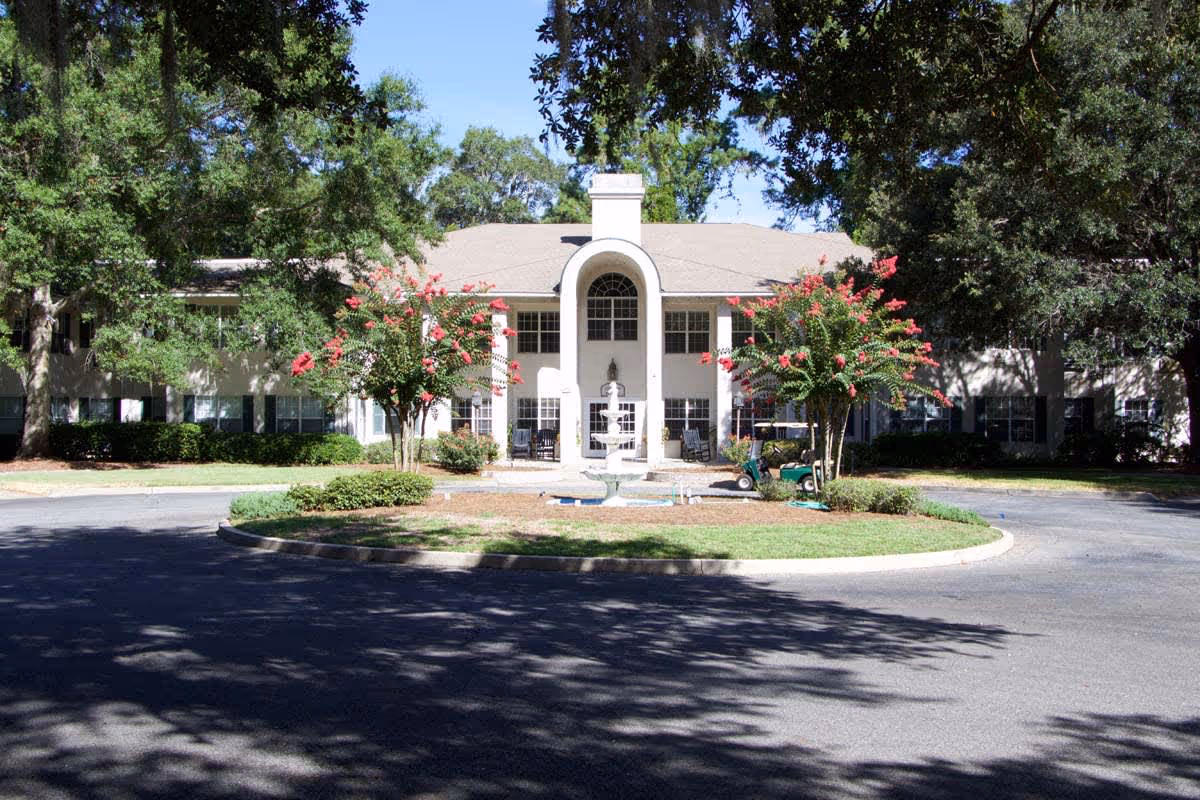 Front exterior view of a two-story retirement living facility with a circular driveway, a central fountain, and flowering trees. The building is surrounded by large trees and greenery under a clear blue sky.