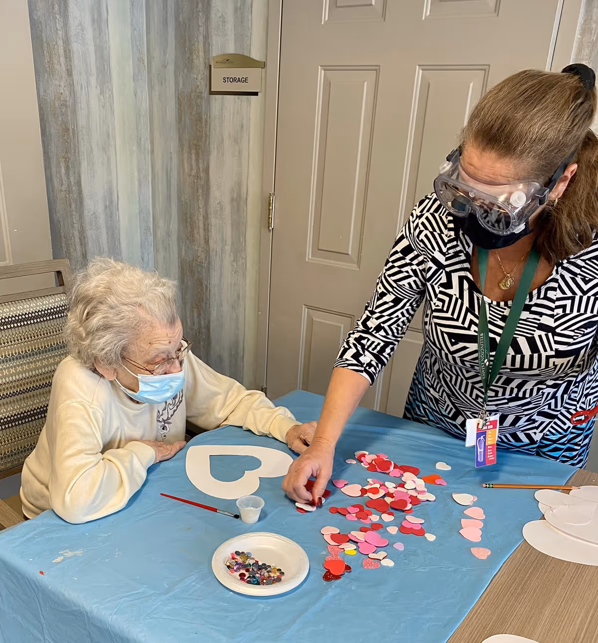An elderly woman wearing a face mask sits at a table covered with a blue tablecloth, watching a woman wearing protective goggles and a face mask arrange colorful heart-shaped cutouts and craft supplies on the table in a room with a door labeled 'STORAGE'.