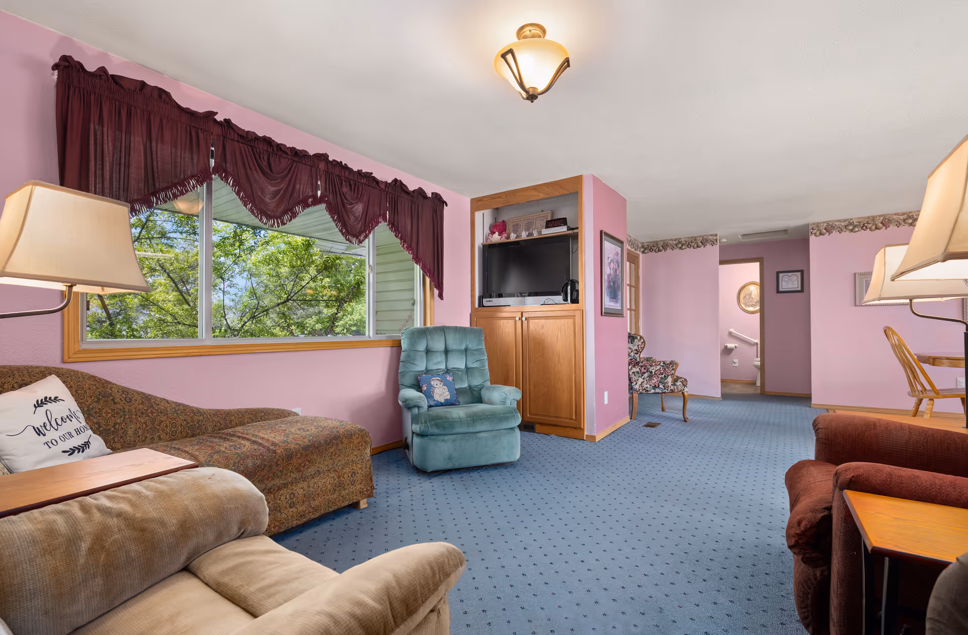 Cozy pink-walled senior living common area with seating, TV, and large window views.
