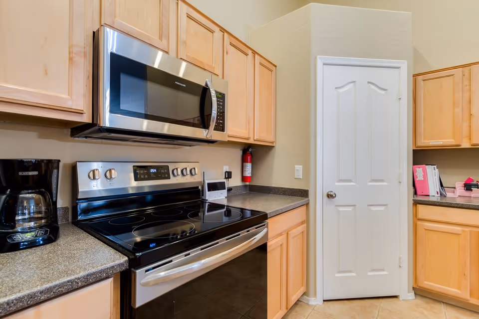 A modern kitchen with light wood cabinets, a stainless steel microwave mounted above a black electric stove with a glass cooktop, a black coffee maker on the countertop, and a white door in the corner. The countertops are dark gray and the walls are beige.