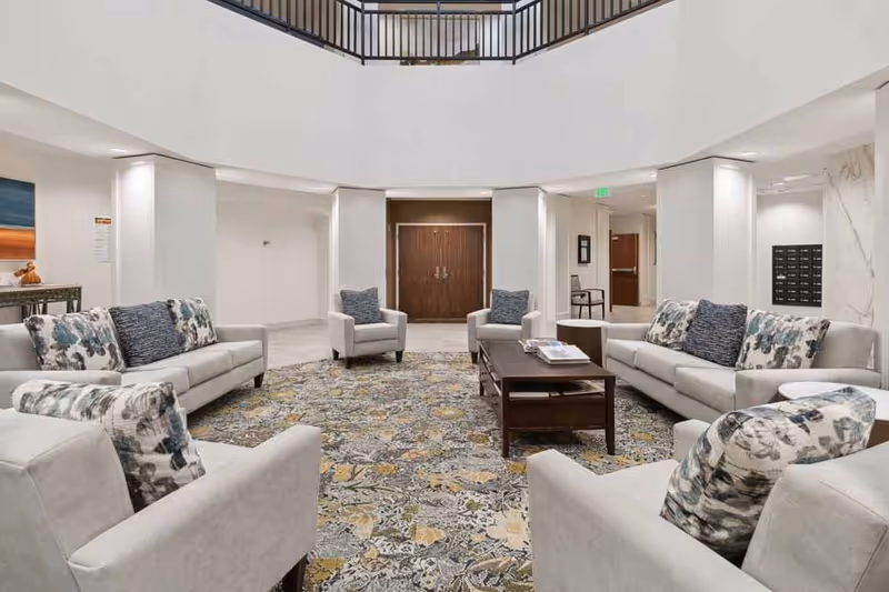 A spacious and well-lit common area with beige sofas and armchairs arranged around a dark wooden coffee table on a patterned carpet. The room features white walls, a high ceiling with a balcony railing above, and double wooden doors in the background.