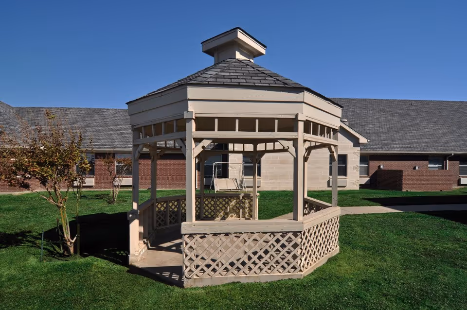 Wooden gazebo with lattice railing sits on a grassy lawn in front of a brick building.