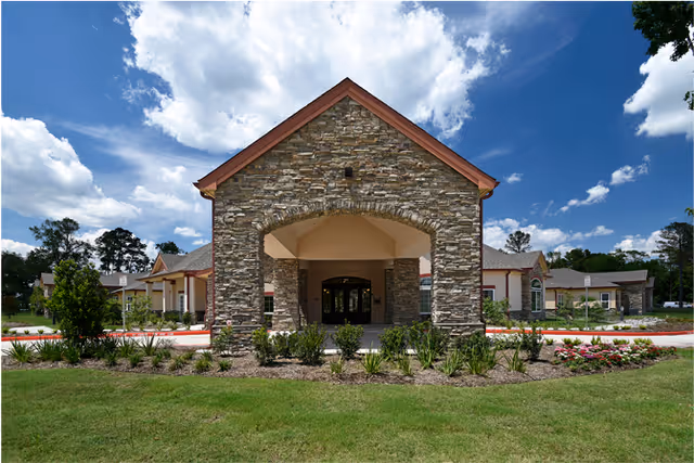 Front exterior view of a senior living community building with a stone facade entrance, landscaped garden beds, and a clear blue sky with some clouds.