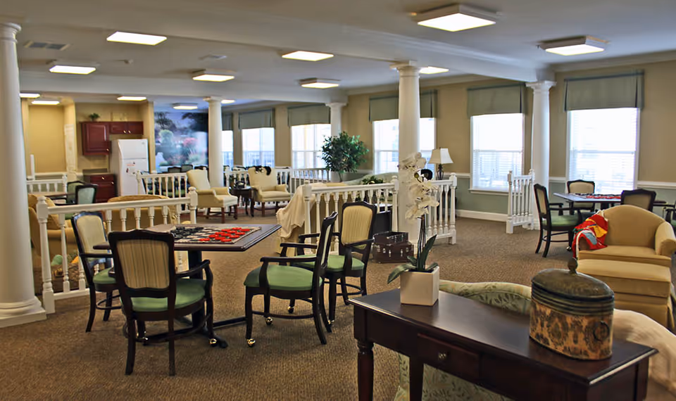 A spacious senior living common area with multiple seating arrangements including chairs and small tables. There are columns and large windows with blinds letting in natural light. The room has a carpeted floor, a few plants, and a cozy atmosphere with a mix of armchairs and tables, some set up for board games.