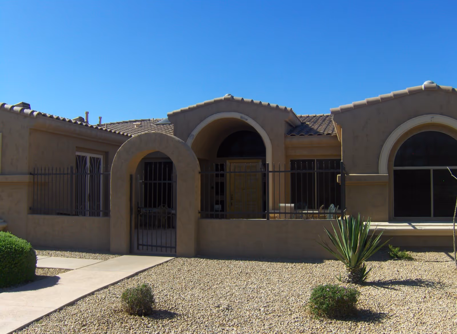 Exterior view of a single-story building with a beige stucco finish and a tiled roof under a clear blue sky. The building features arched windows and an arched gated entrance leading to a small patio area. The front yard is landscaped with gravel and a few small bushes and desert plants.