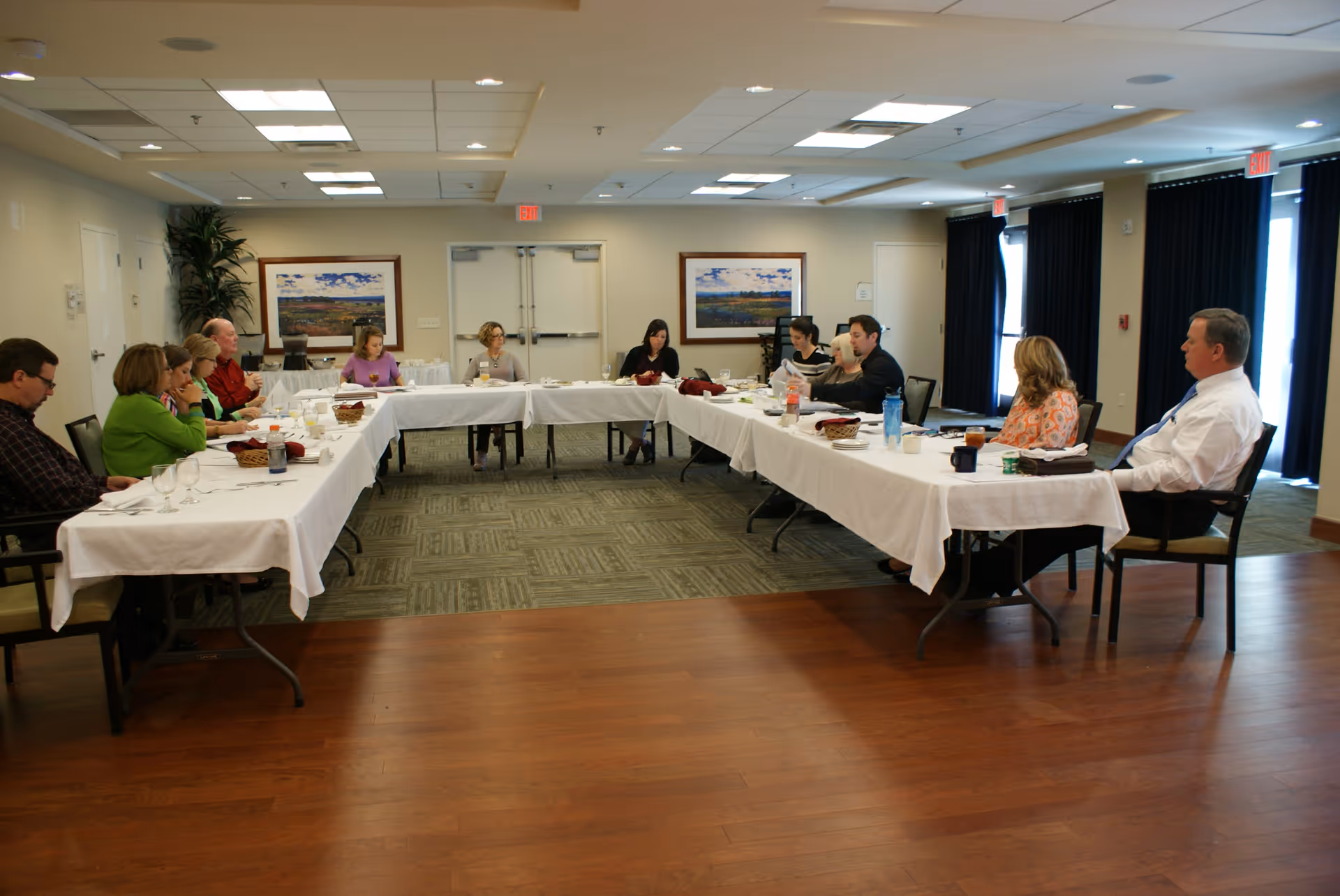 A group of people seated around a large U-shaped table covered with white tablecloths in a conference or meeting room. The room has carpet and wood flooring, framed landscape paintings on the walls, and large windows with dark curtains. The attendees appear to be engaged in a discussion or meeting.