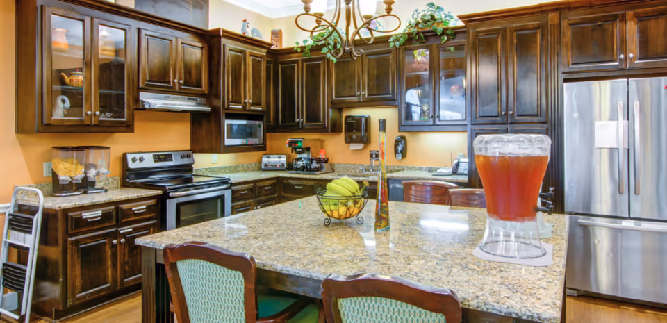 Bright communal kitchen with a large granite island, dark wood cabinets, stainless steel appliances, a fruit bowl and a beverage dispenser.