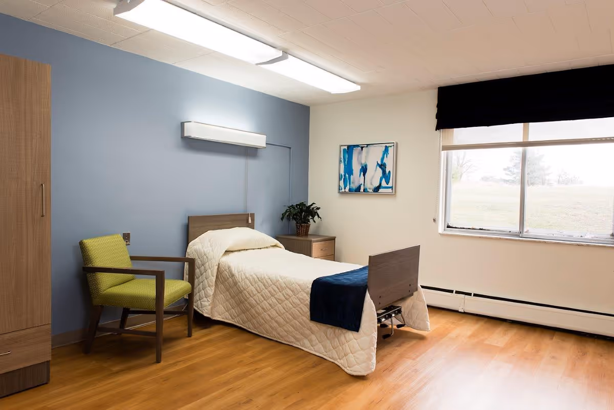 A simple, clean bedroom in a senior living facility featuring a single bed with a white quilted bedspread and a dark blue throw. The room has light wooden flooring, a green cushioned chair with wooden arms, a wooden wardrobe, and a small wooden nightstand with a potted plant. There is a large window with a black valance and a white roller shade, and a blue and white abstract painting on the wall.