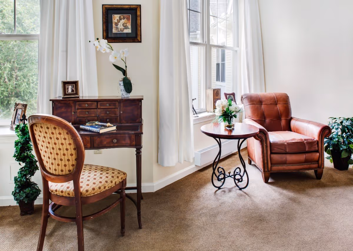 A cozy sitting area with a vintage wooden desk and patterned chair on the left, a small round table with a flower vase in the center, and a brown leather armchair on the right. The room has large windows with white curtains, beige carpet, and some green plants.