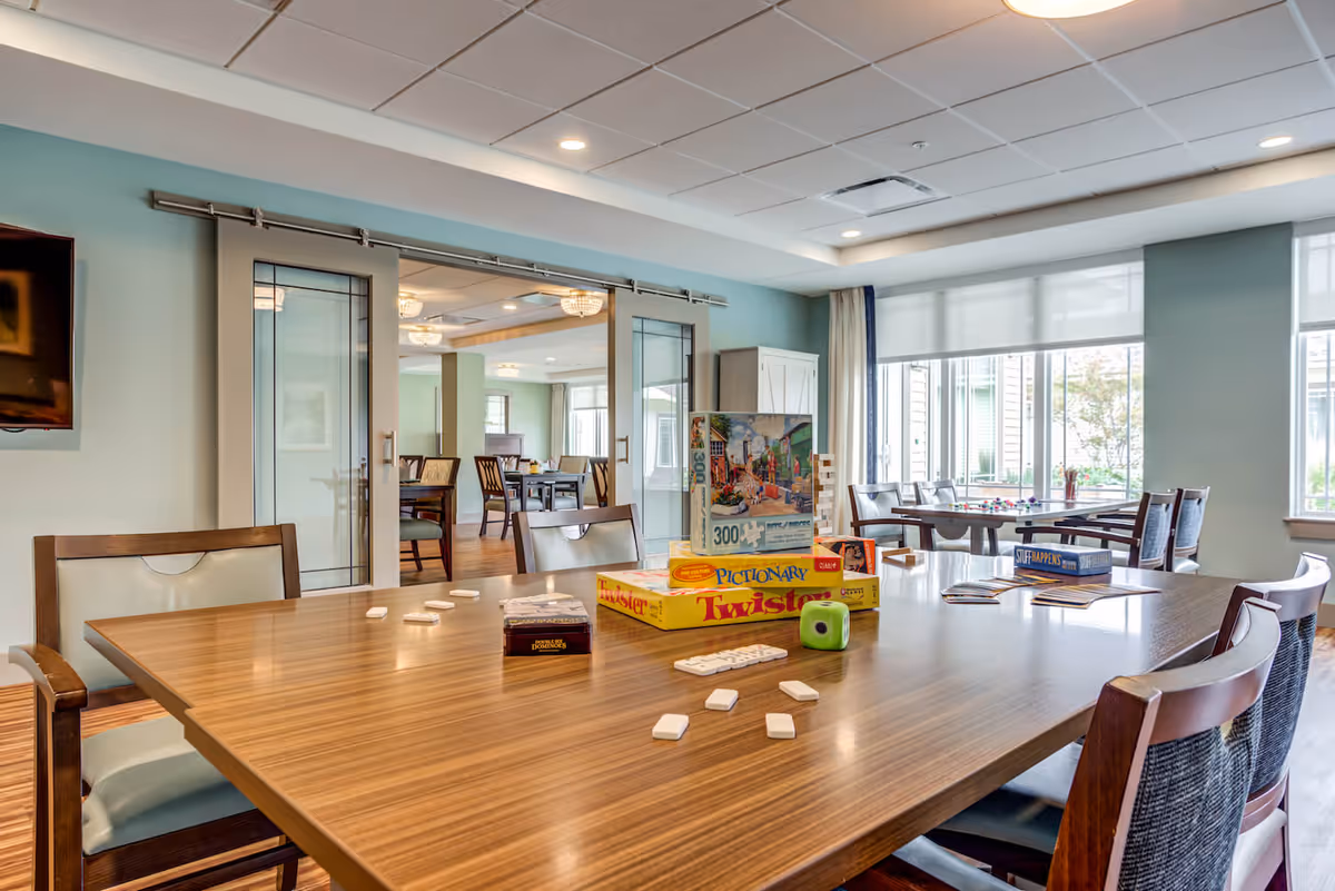 Bright communal dining/activity room with a large table covered in board games and chairs, sliding glass doors, and windows letting in natural light.