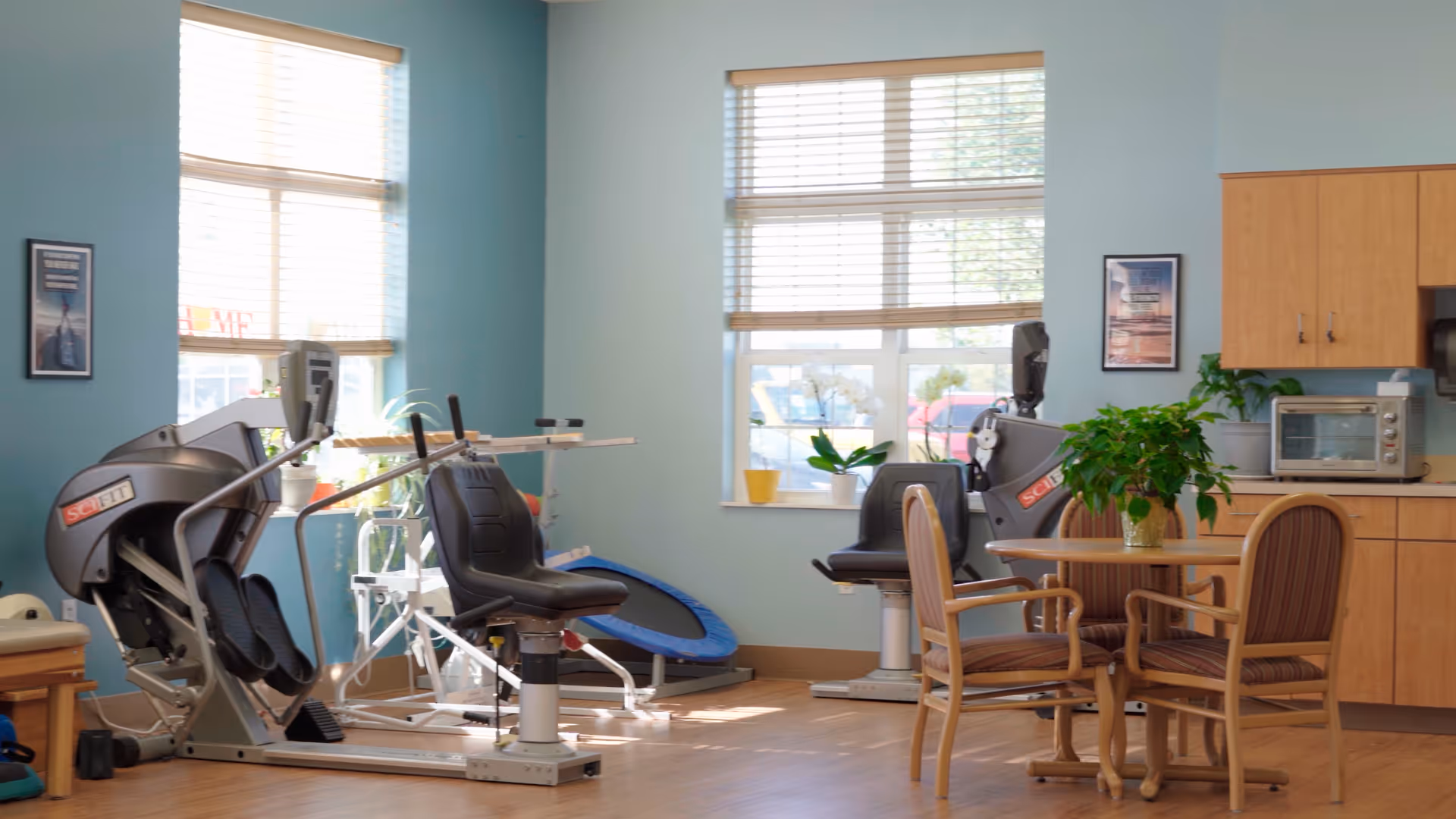 A bright room with exercise equipment including two seated elliptical machines and a small trampoline, alongside a round wooden table with four striped cushioned chairs and a green potted plant on top. The room has light blue walls, two large windows with blinds, wooden cabinets, and a countertop with a microwave and other items.