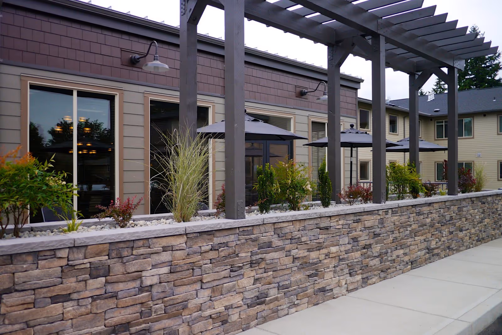 Outdoor patio area of a senior living facility with a stone wall planter containing various shrubs and plants. There are large windows on the building behind the planter, and several black patio umbrellas providing shade. A wooden pergola structure extends over the patio area.