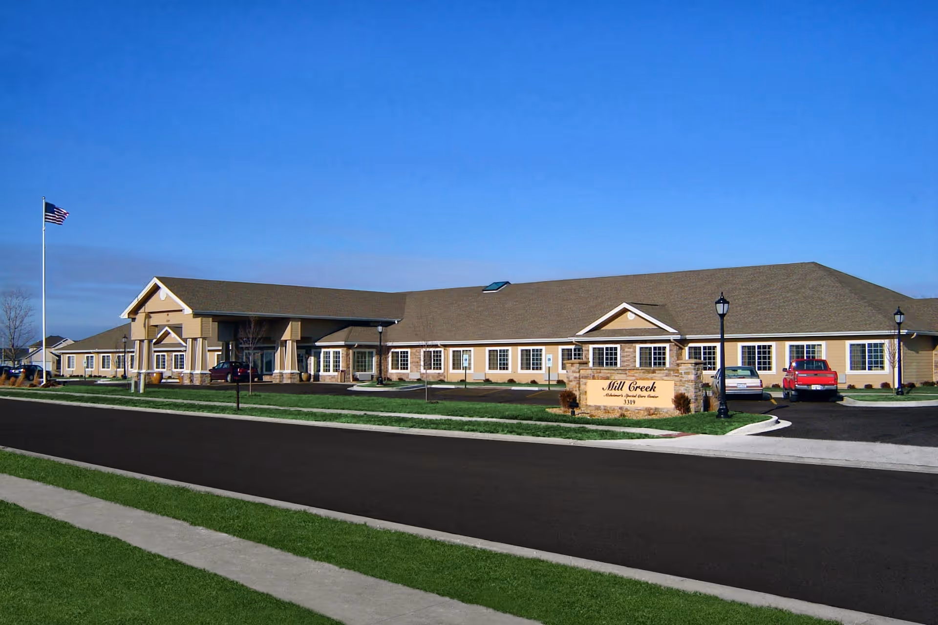 Front view of the single-story Mill Creek Alzheimer's Special Care Center building with a driveway, parked cars, flagpole and lawn under a clear blue sky.
