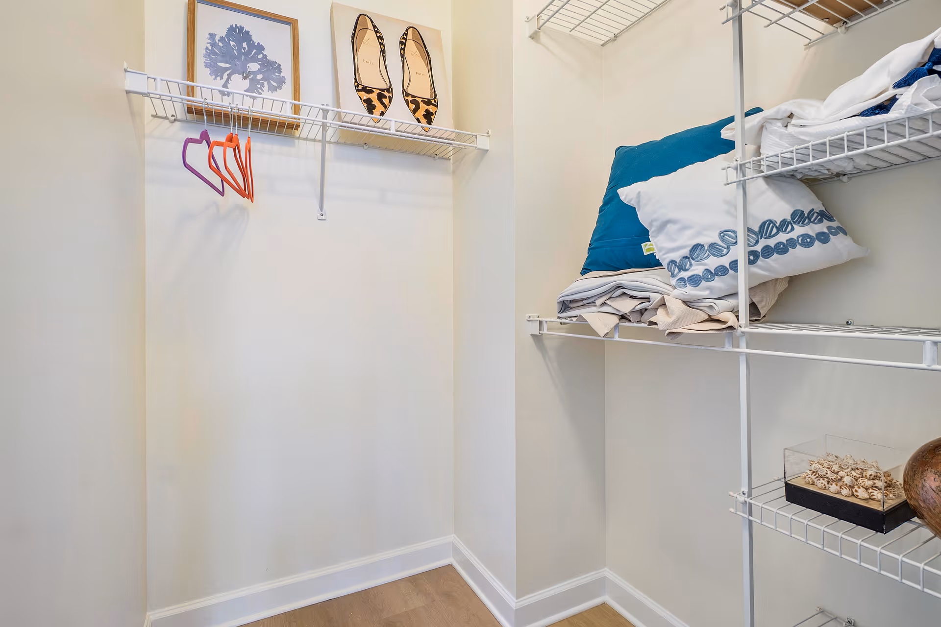 Small walk-in closet with white wire shelving, a few colorful hangers, folded linens and decorative pillows.