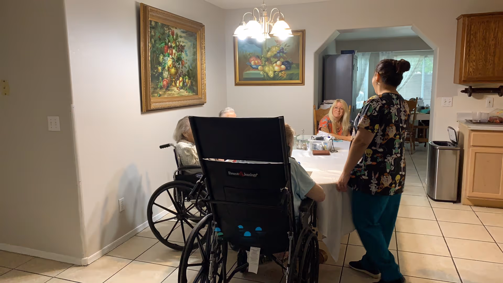 A home dining area with a table where two wheelchair users sit, a seated woman across from them, and a caregiver standing nearby.