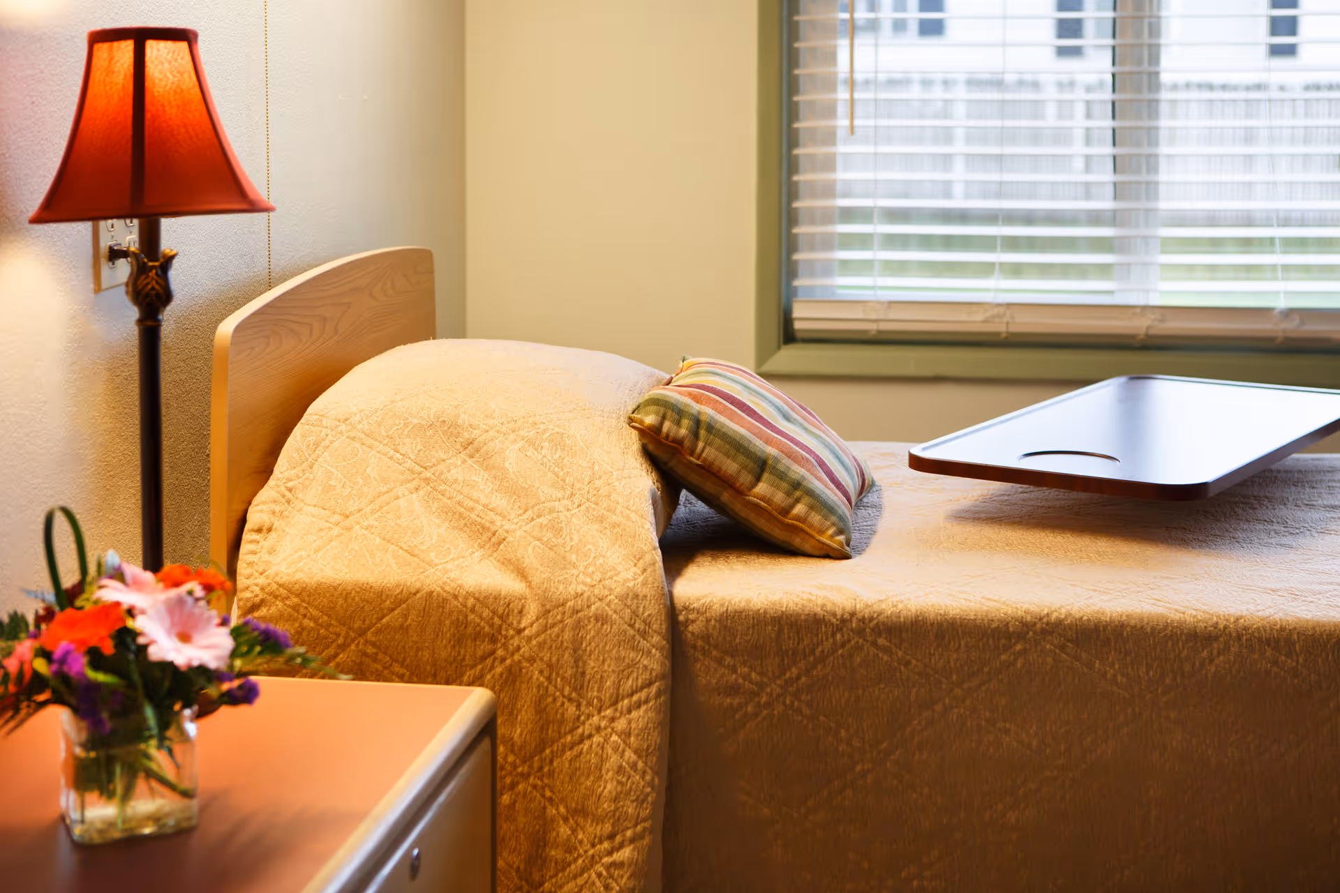 A cozy bedroom with a single bed covered in a beige quilt. There is a striped pillow and a wooden overbed table on the bed. A red lampshade on a floor lamp is beside the bed, and a small vase with colorful flowers sits on a nearby surface. A window with white blinds is in the background.