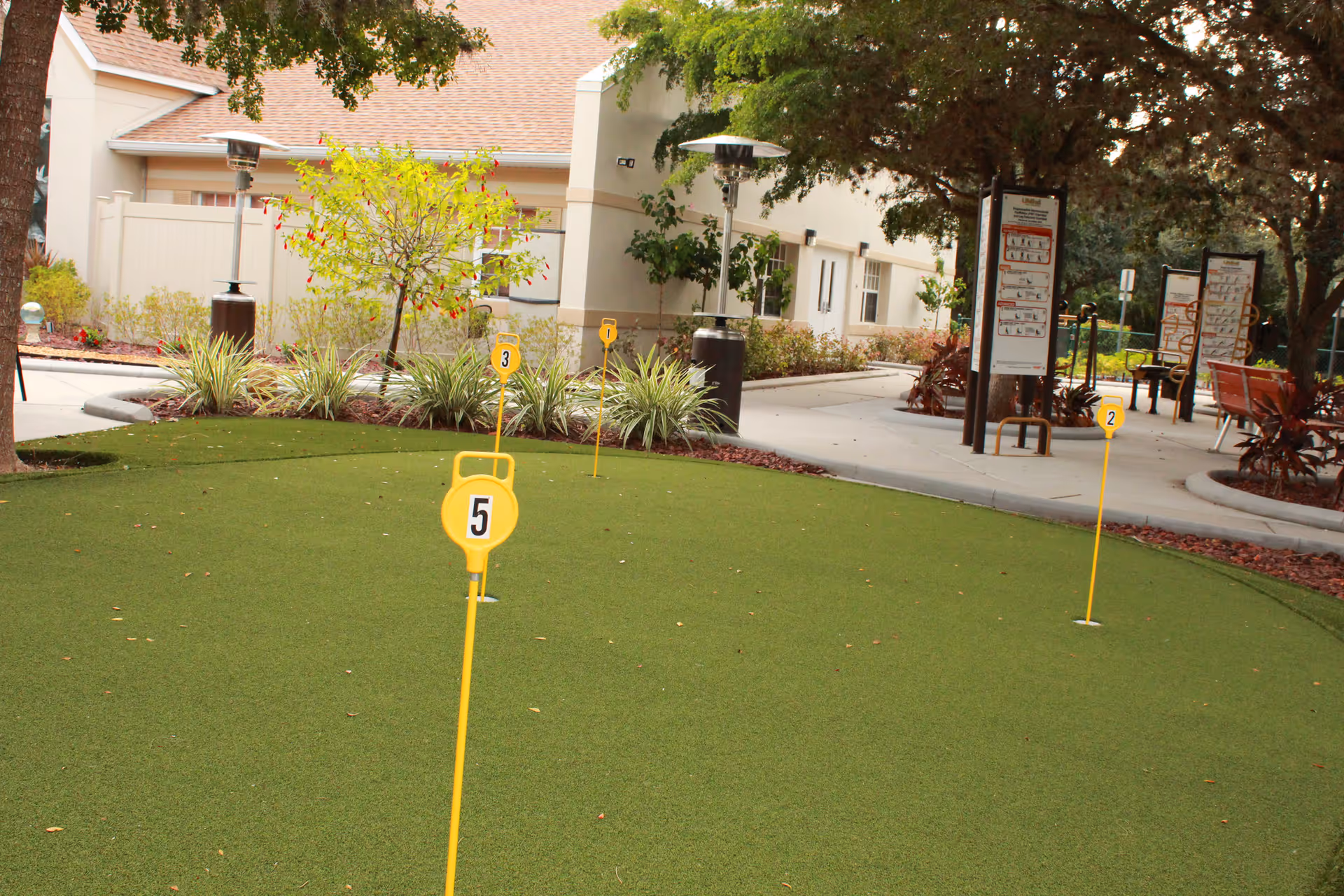 Outdoor putting green area with numbered yellow markers on artificial grass, surrounded by plants, trees, and a building in the background. There are also outdoor exercise equipment and informational signs visible near the paved walkway.