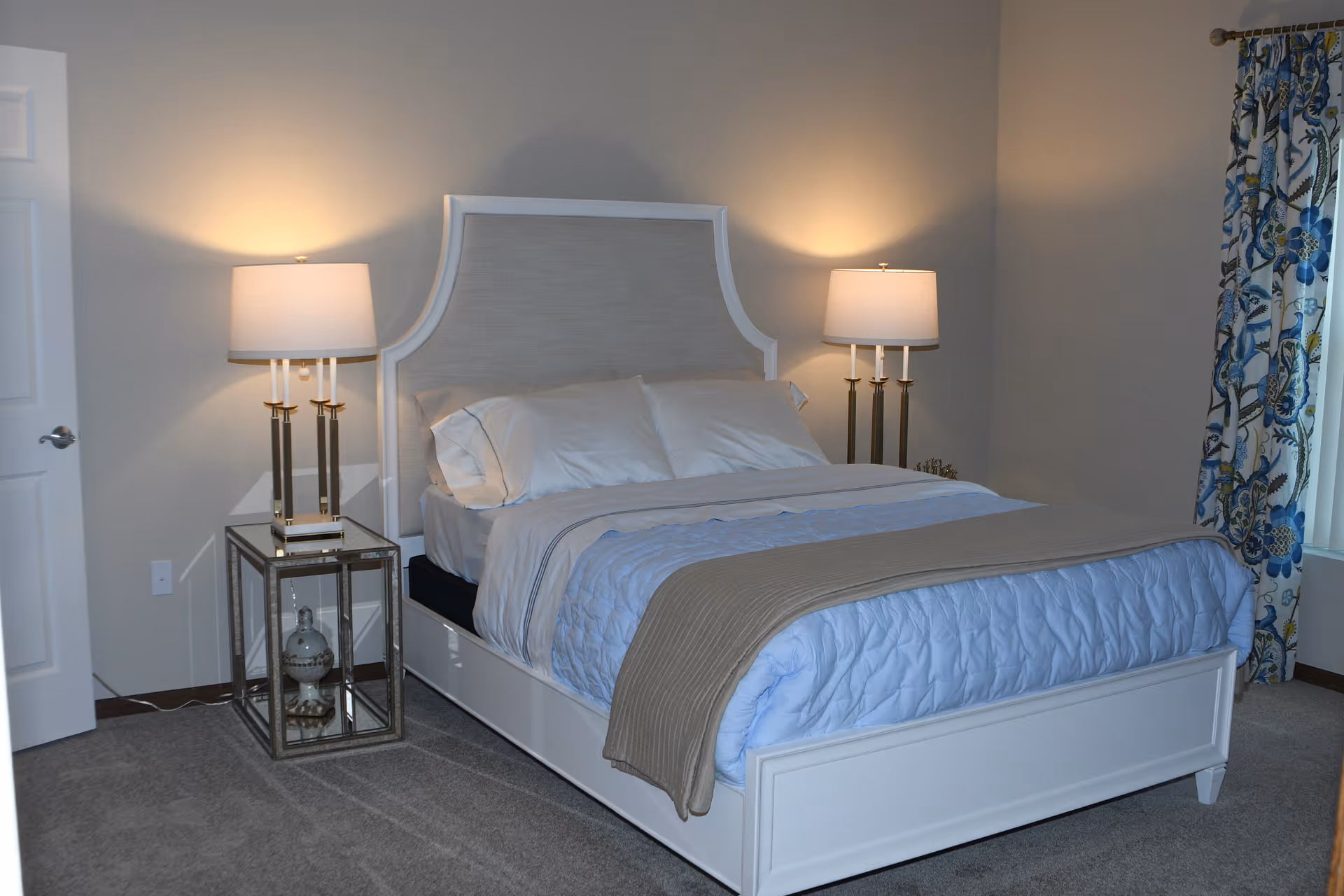 A neatly made bed with a white frame and upholstered headboard in a bedroom. The bed has white pillows, a light blue quilt, and a beige throw blanket. There are two matching bedside tables with glass sides, each holding a lamp with a white shade. A window with floral curtains is visible on the right side of the room, and a closed white door is on the left.