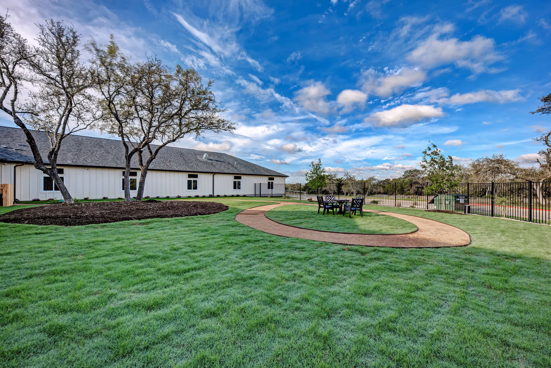 Well-maintained grassy courtyard with a circular walking path, outdoor seating, trees, and a low white building under a blue sky.