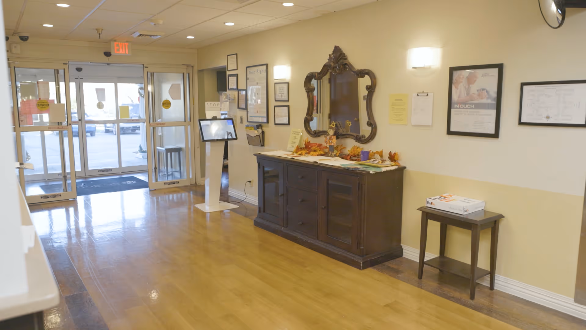 Interior view of the entrance area of a facility with glass double doors leading outside. There is a wooden sideboard with a decorative mirror above it on the right wall, decorated with autumn-themed items. A small table with a FedEx box is also visible. Various framed documents and notices are hung on the walls. The floor is wooden and shiny, and there is an exit sign above the doors.