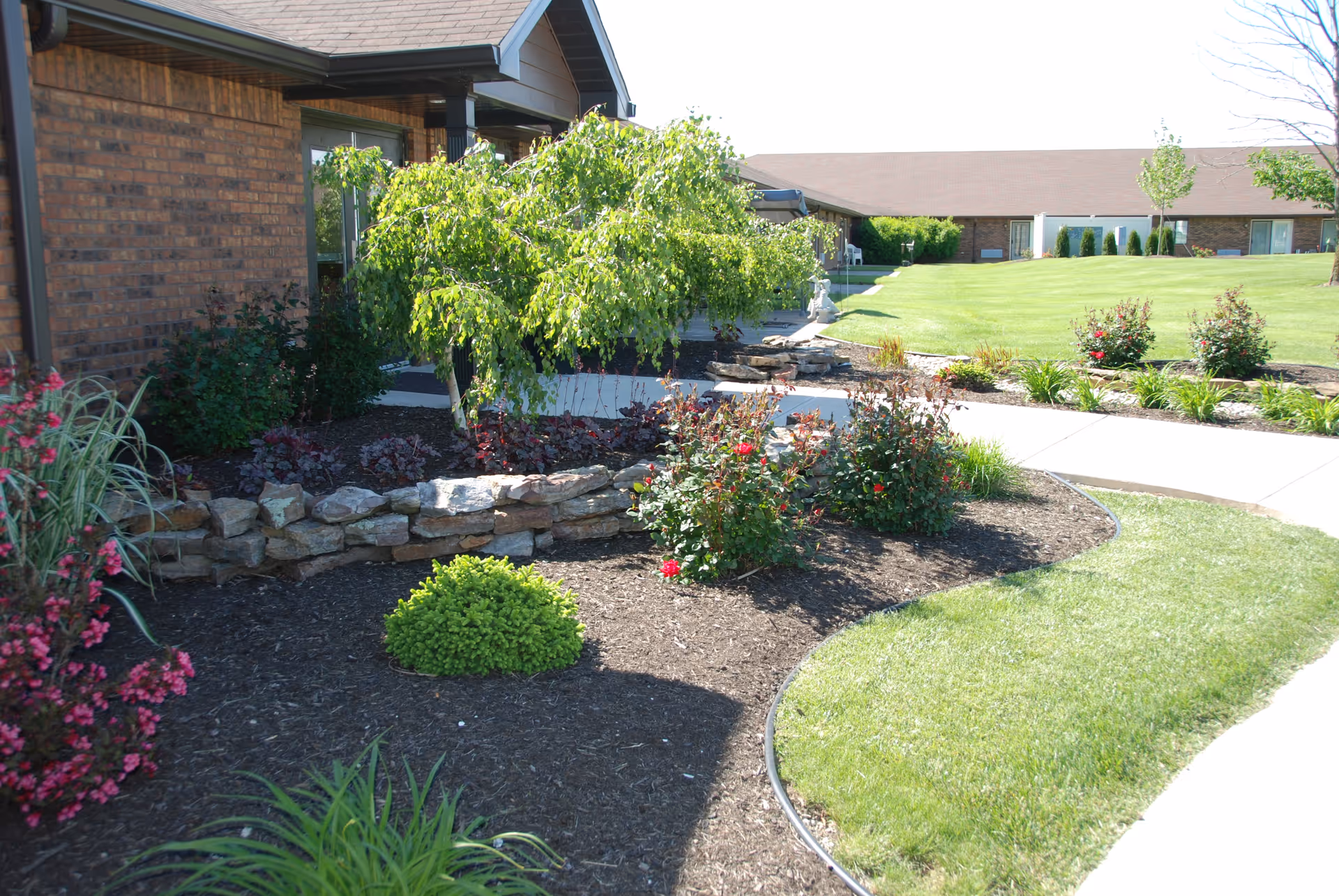 Landscaped front lawn and walkway with shrubs and flowers beside the brick exterior of a senior living facility.