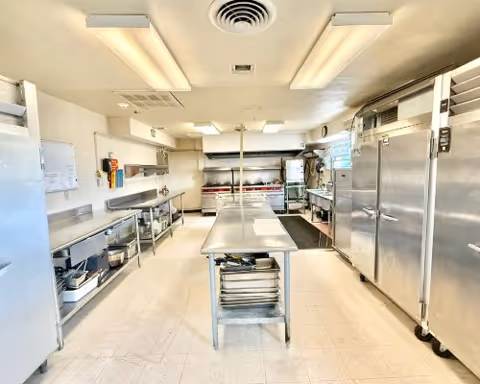 A clean, well-lit commercial kitchen with stainless steel appliances and work surfaces. There are large refrigerators on the right, a central stainless steel prep table with trays underneath, and various kitchen equipment along the walls including ovens, sinks, and shelves with utensils and containers.