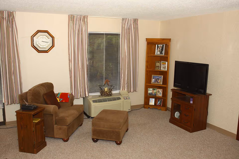 A cozy living room with a brown armchair and matching ottoman, a wooden side table with a radio, a wooden corner shelf displaying framed photos, a flat-screen TV on a wooden TV stand, beige carpet, and a window with striped curtains and blinds.