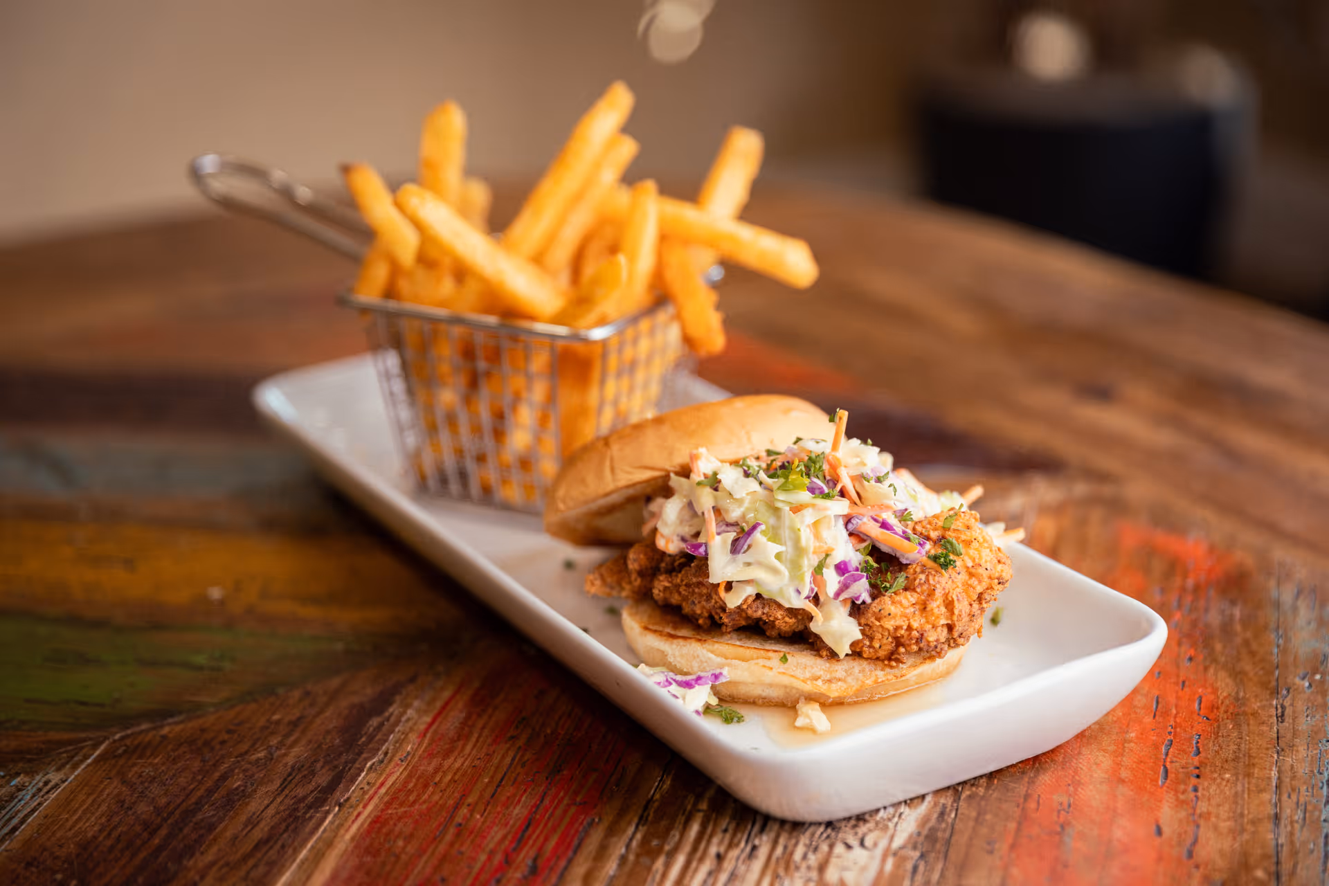 A plate with a fried chicken sandwich topped with coleslaw and a side of French fries served in a small metal basket on a rustic wooden table.