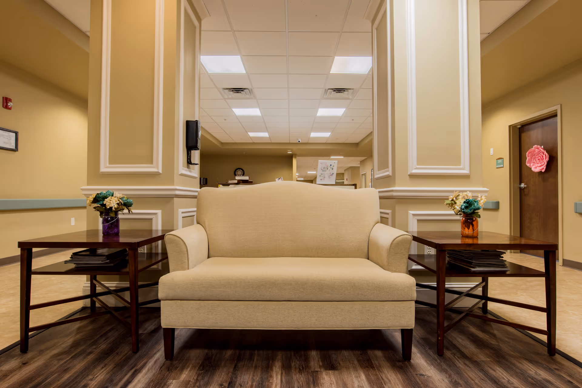 Beige loveseat centered between two wooden side tables with flower arrangements in a senior facility lobby hallway.