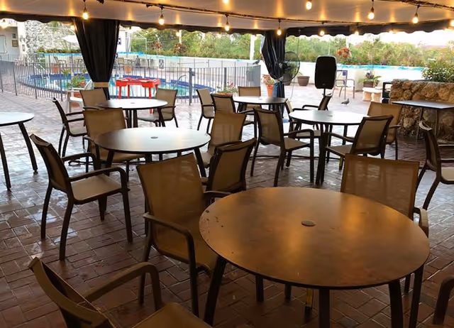 Outdoor covered seating area with multiple round tables and chairs arranged on a brick floor. String lights hang from the ceiling, and a fenced pool area is visible in the background along with some greenery.