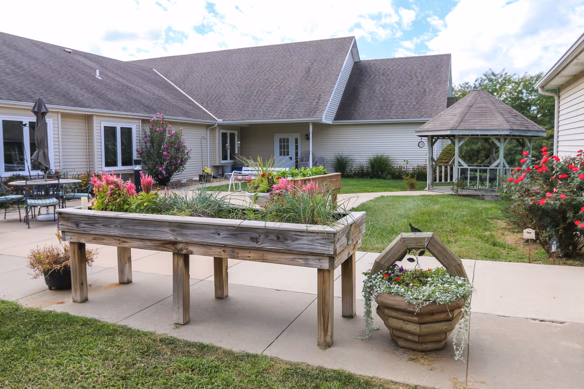 Courtyard with raised wooden planters, potted flowers, patio seating and a gazebo outside a single-story senior living building.