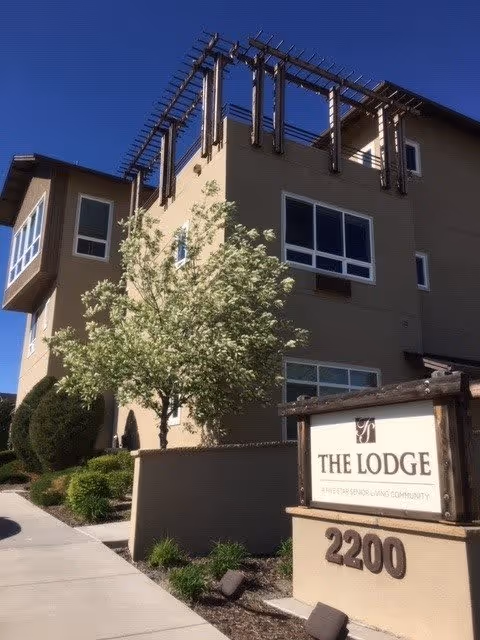Exterior view of The Lodge Assisted Living And Memory Care Community building with beige walls, multiple windows, and a tree in front. A sign near the sidewalk displays the facility name and address number 2200.