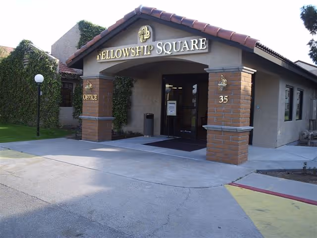 Entrance of the Fellowship Square building with a tiled gabled canopy, brick columns, glass doors, and visible 'FELLOWSHIP SQUARE' signage.