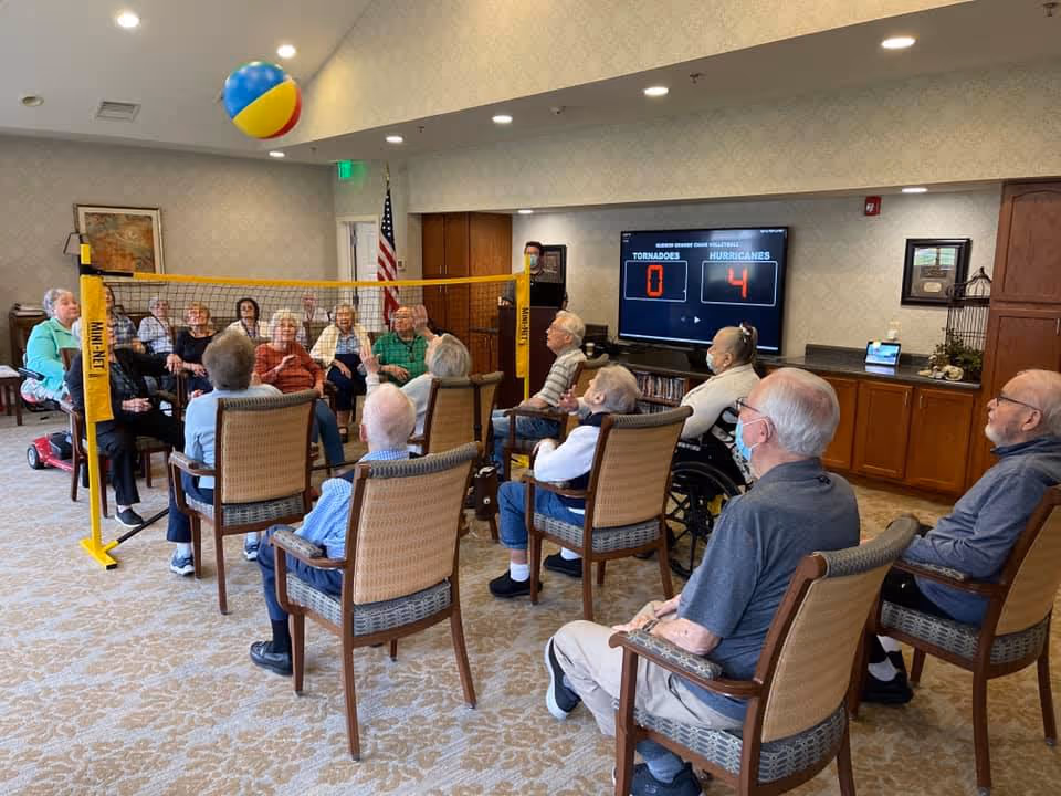 A group of elderly people seated in chairs arranged in a circle in a common room, playing a seated volleyball game with a beach ball over a mini net. A large screen in the background shows the score with Hurricanes leading Tornadoes 4 to 0. The room has carpeted floors, wooden cabinets, and an American flag in the corner.