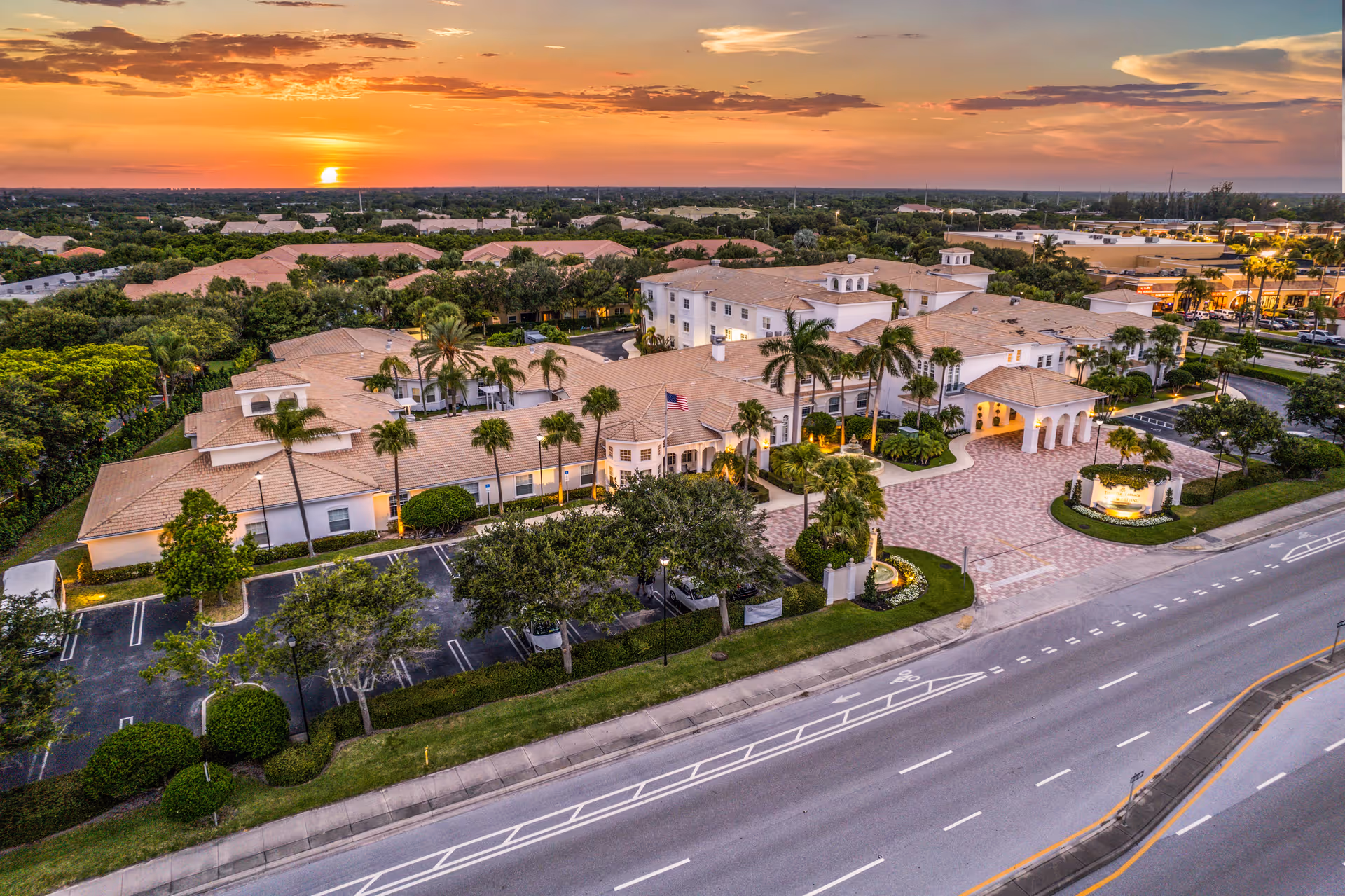 Aerial view of Tequesta Terrace senior living facility at sunset, showing a large building complex with beige roofs, surrounded by palm trees and greenery, with a paved entrance and parking areas visible along a main road.