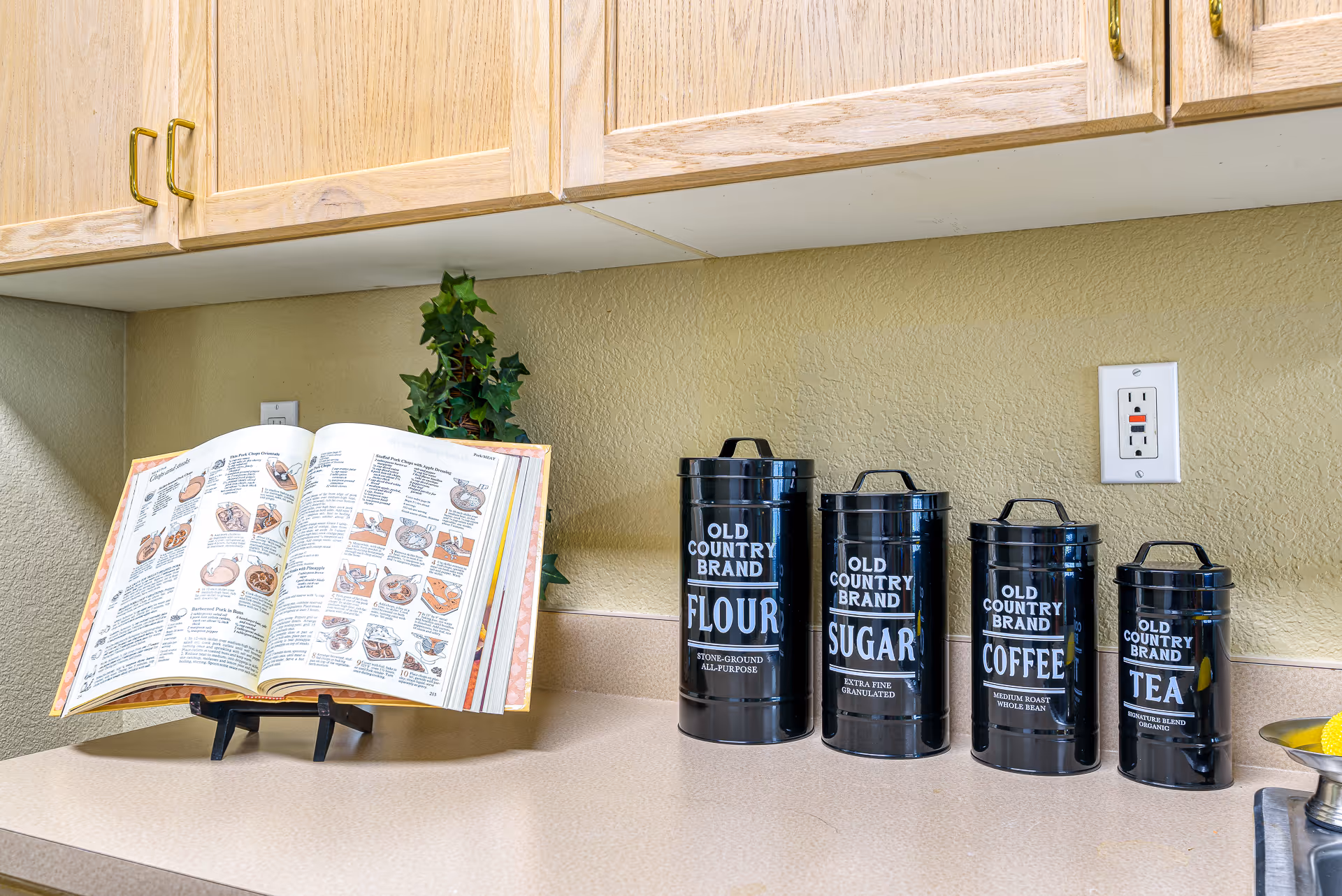 A kitchen countertop with four black canisters labeled Old Country Brand Flour, Sugar, Coffee, and Tea. An open cookbook on a stand is placed on the left side of the counter, and there is a small green plant behind the cookbook. Light wood cabinets are mounted above the countertop, and a power outlet is visible on the wall.