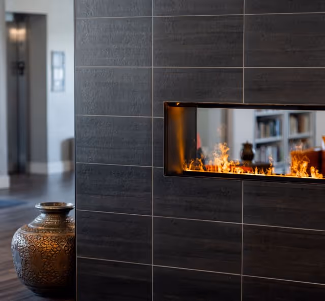 A dark tiled interior wall with a rectangular built-in fireplace showing flames and a decorative ornate vase placed beside it.