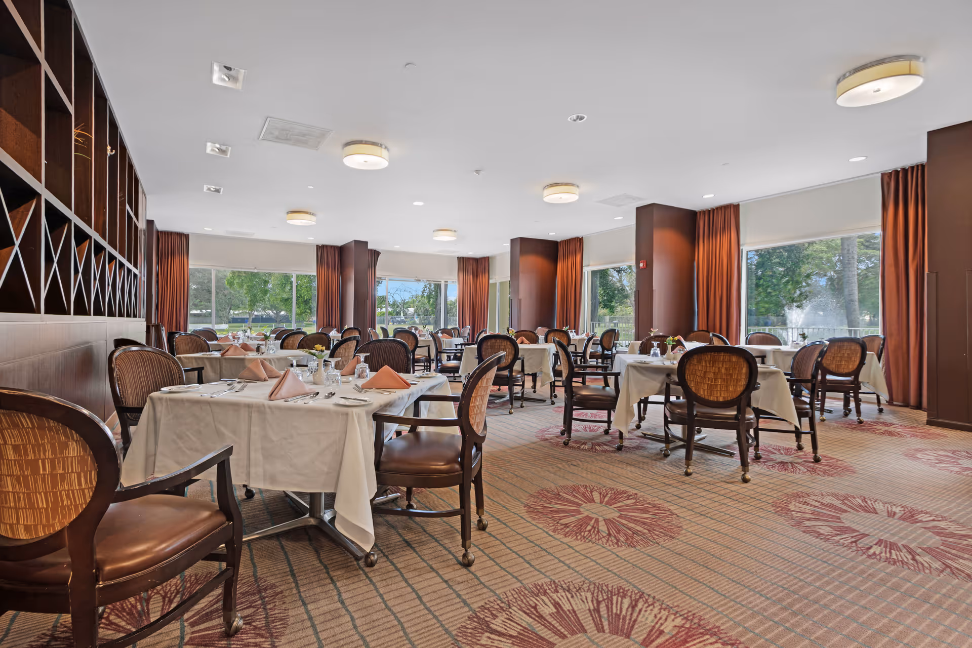 A spacious dining room with multiple tables covered in white tablecloths, each set with plates, glasses, and folded napkins. The room features large windows with brown curtains, allowing natural light to brighten the space. The carpet has a floral pattern, and wooden chairs with cushioned seats surround the tables.