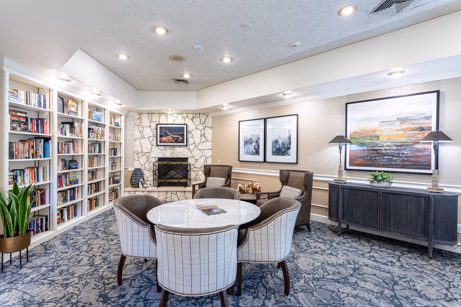 Cozy communal lounge with a round marble table surrounded by upholstered chairs, built-in bookshelves, a stone fireplace, and framed artwork.