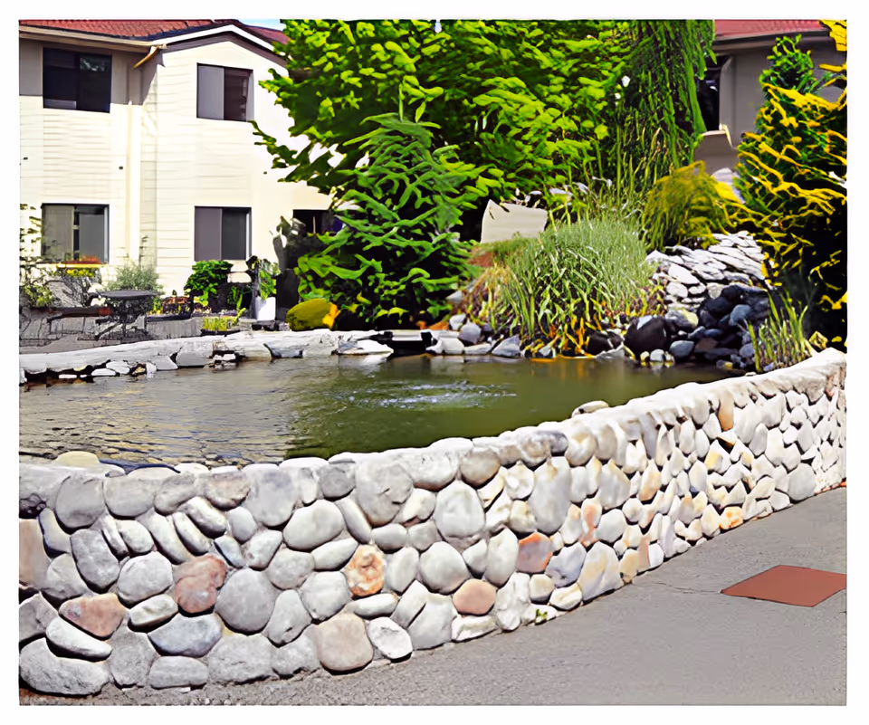A landscaped outdoor area at Mountain Glen Retirement and Assisted Living featuring a stone wall surrounding a pond with water, lush green plants, and trees. In the background, there is a two-story building with windows and a red-tiled roof.
