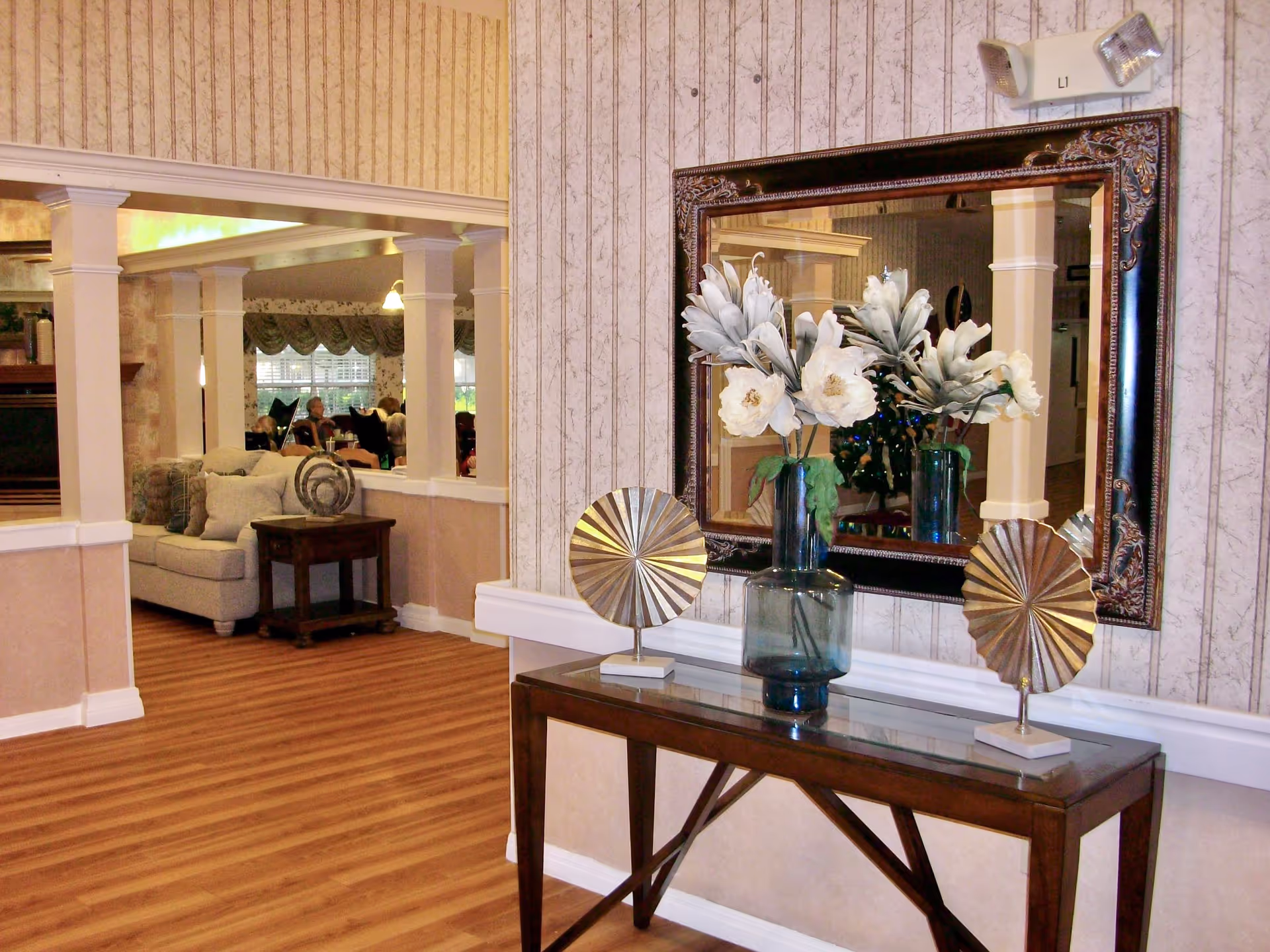 Interior view of a senior living facility showing a wooden console table with decorative items including a vase with white flowers and two metallic fan-shaped sculptures. Above the table is a large ornate mirror reflecting part of the room. The background shows a seating area with sofas, a side table, and people sitting near windows with curtains.