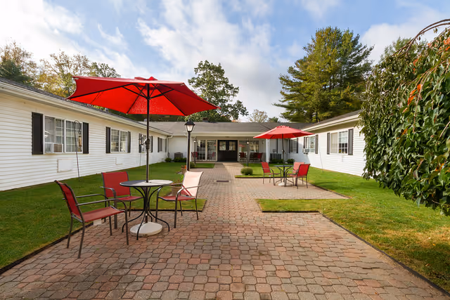 Outdoor courtyard with a brick paver walkway, red umbrellas and patio tables between single-story white building wings.