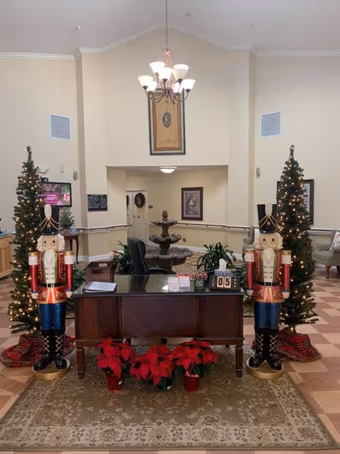 Reception area decorated for the holidays with two large nutcracker statues on either side of a wooden desk. The desk has poinsettia plants in front of it and a small calendar showing the date. Behind the desk is a fountain and some framed artwork on the walls. Two decorated Christmas trees with lights flank the nutcrackers. The room has a high ceiling with a chandelier and beige walls.