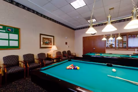 Interior game room with a pool table, racked balls and cues, seating along the wall, and hanging lights.