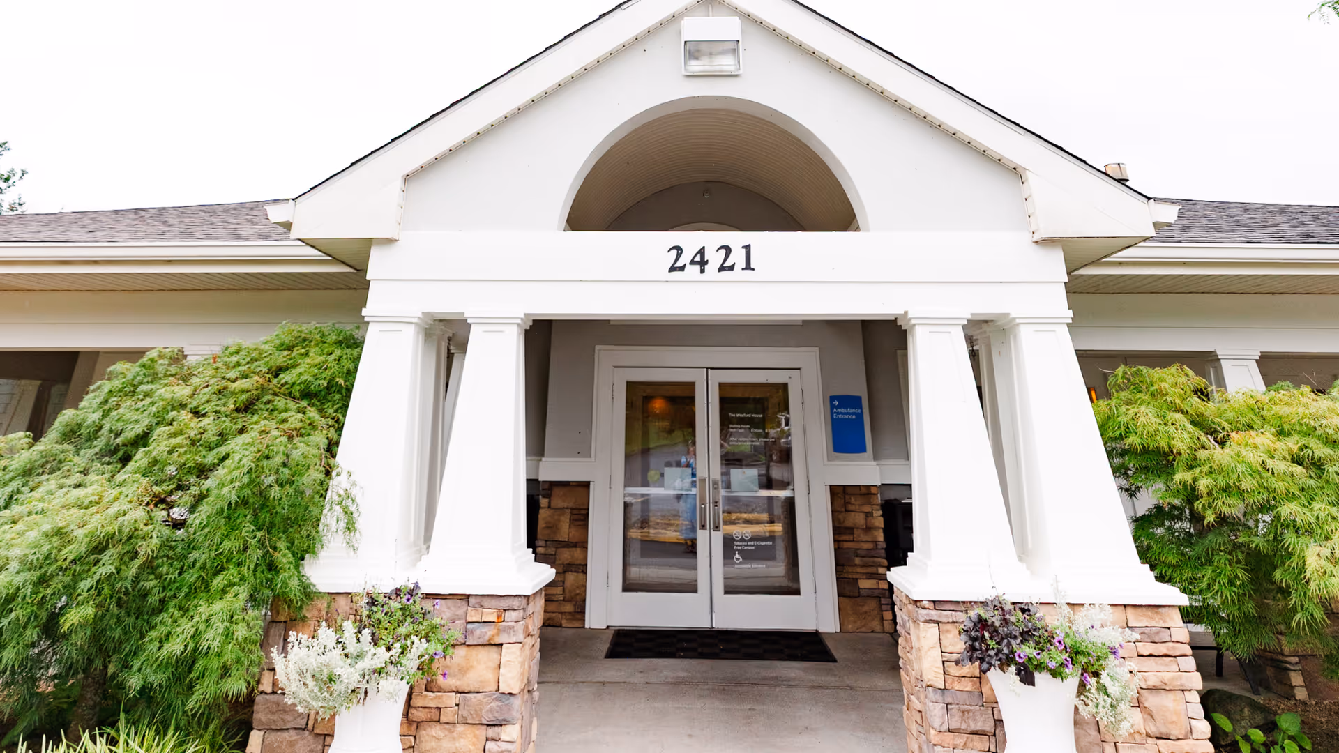 Front entrance of a building with white pillars and stone accents, featuring double glass doors under a peaked roof with the number 2421 displayed above. Green shrubs and plants flank the entrance.