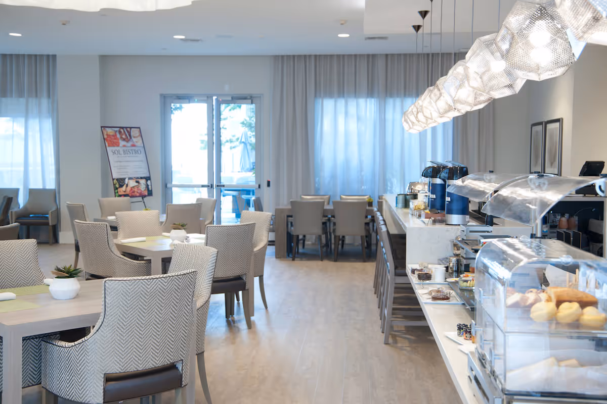 Bright dining room with tables and upholstered chairs and a buffet counter displaying pastries and beverage dispensers.