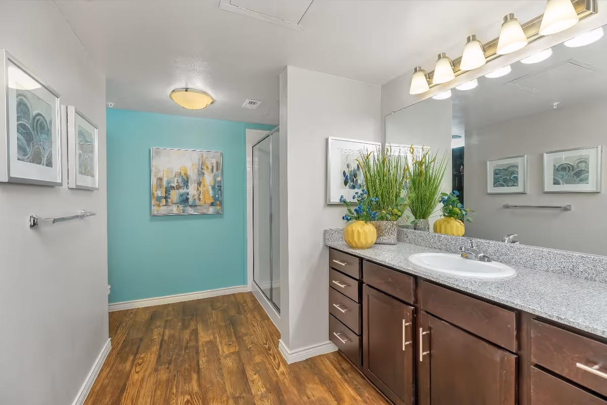 Bathroom with wood-look flooring, a teal accent wall and glass shower on the left, and a long vanity with granite countertop, sink and decorative plants on the right.